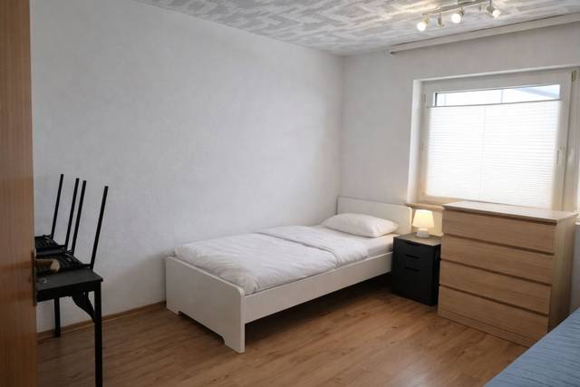 Einzelbett mit schwarzem Nachttisch und Kommode vor dem Fenster in einem Monteurzimmer in Frankfurt am Main